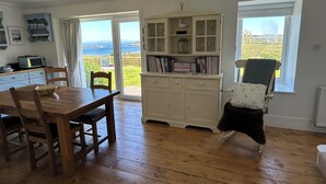 Een eetruimte met een houten tafel en stoelen, een wit kastje met glazen deuren en uitzicht op zee door een groot raam.