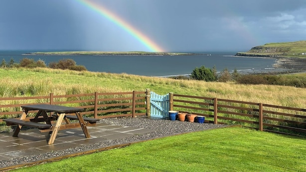 Picknickplaats met een houten tafel en banken, met uitzicht op een waterpartij, een regenboog en een verre kustlijn.