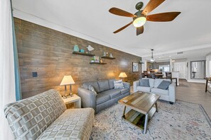 Sala de estar con dos sofás, una mesa de centro y un ventilador de techo. Se ve una cocina al fondo.