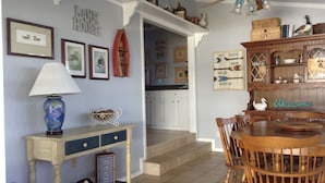 A dining area with a wooden table and chairs, a decorative lamp, framed pictures on the wall, and a 'Welcome' sign.