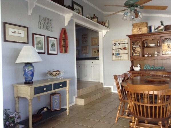 A dining area with a wooden table and chairs, a decorative lamp, framed pictures on the wall, and a 'Welcome' sign.