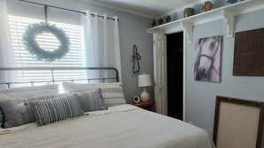 A bedroom with a bed, a bedside table with a lamp, a door, and a wall with a wreath and a painting.