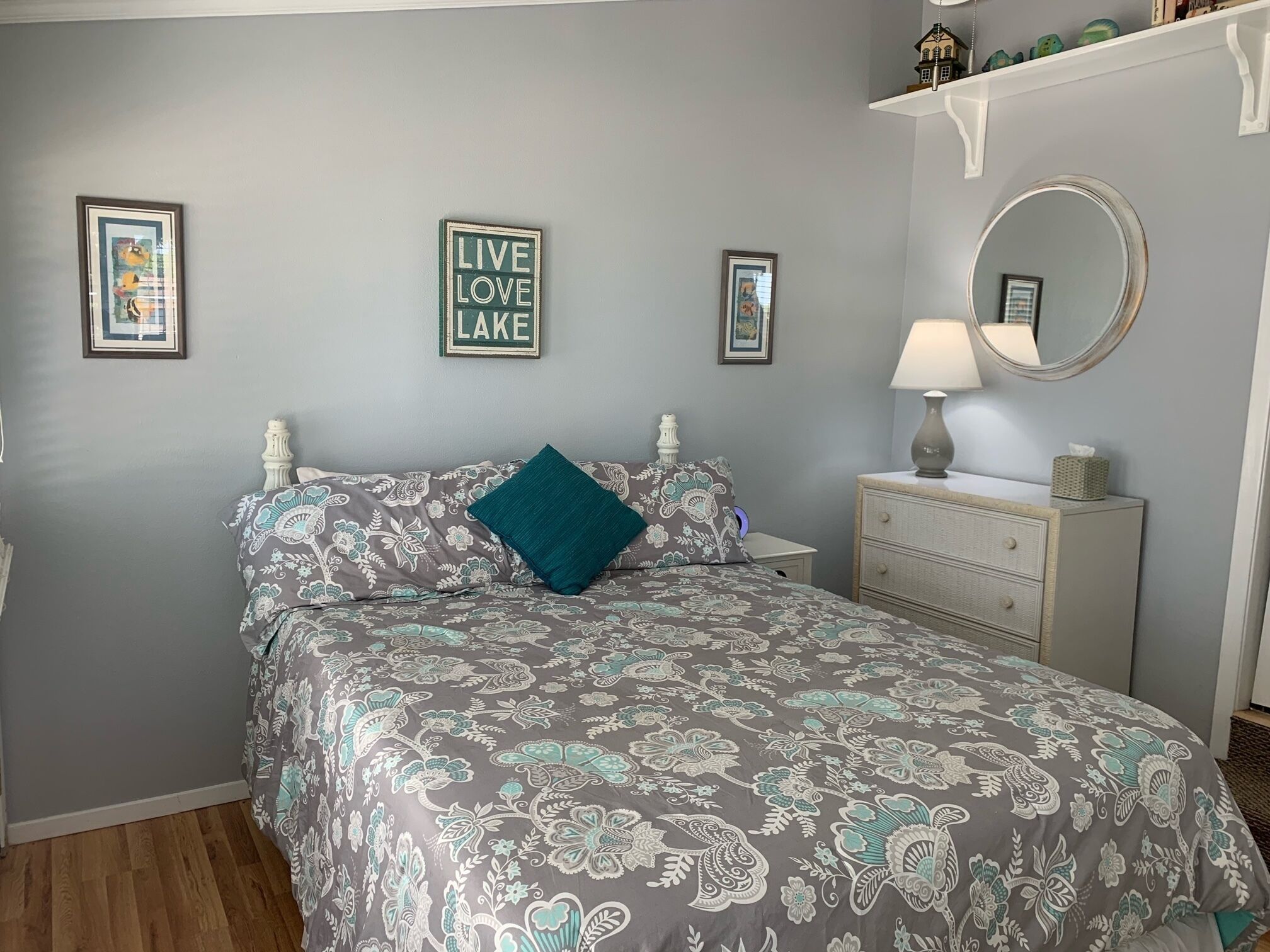 Un dormitorio con una colcha estampada de flores, una mesita de noche con lámpara, un aparador y dos cuadros en la pared.