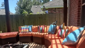 Un área de patio techada con un sofá naranja curvo, varios cojines de colores, una mesita y un brasero.