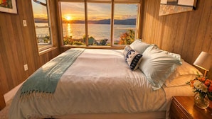 A bedroom with a large window, a bed, a nightstand, and a vase with flowers.