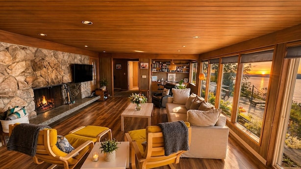 A modern living room with a stone fireplace, wooden flooring, yellow chairs, a sofa, and large windows offering a view of the sunset.