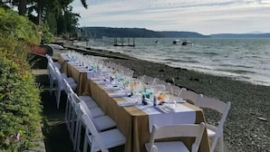 A long table set up on a pebbled beach with chairs, overlooking a body of water and distant hills.