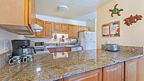 A kitchen with granite countertops, wooden cabinets, a coffee maker, a toaster, and a bulletin board on the wall. - Tropical 2-BR Condo + Pool +  Hot Tub! 
CHECK OUT OUR SPRING 2026 SPECIALS (Kihei)