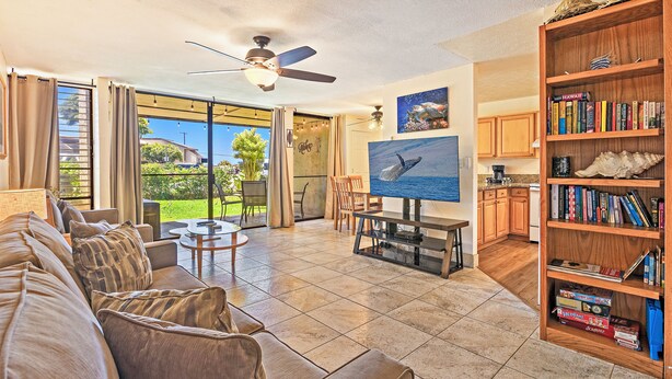 A spacious living room with a large sofa, a dining table, and a bookshelf filled with books. - Tropical 2-BR Condo + Pool +  Hot Tub! 
CHECK OUT OUR SPRING 2026 SPECIALS (Kihei)