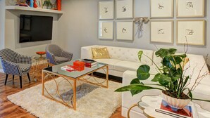 Una sala de estar con un sofá blanco, dos sillones estampados, una mesa de centro de vidrio, un televisor y cuadros en la pared.