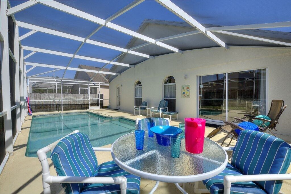 A covered outdoor pool area with lounge chairs and a table, large windows, and a clear view of the pool.
