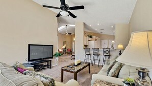 A modern living room with a flat-screen TV, a ceiling fan, and a dining area with bar stools. - Minutes from Disney - Beautiful 3/2 Pool Villa with WiFi in Hampton Lakes  (Davenport)