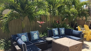 An outdoor seating area with a sectional sofa and chairs, surrounded by lush greenery and a wooden fence.