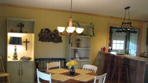 Comedor con mesa de madera y sillas blancas, una araña y área de cocina con fregadero y taburetes de bar.
