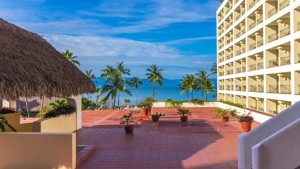A hotel courtyard with a view of the ocean, palm trees, and a thatched-roof structure.