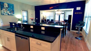 A modern kitchen with a black countertop, white cabinets, and a dishwasher. There is a dining area with a table and chairs visible through an open doorway.