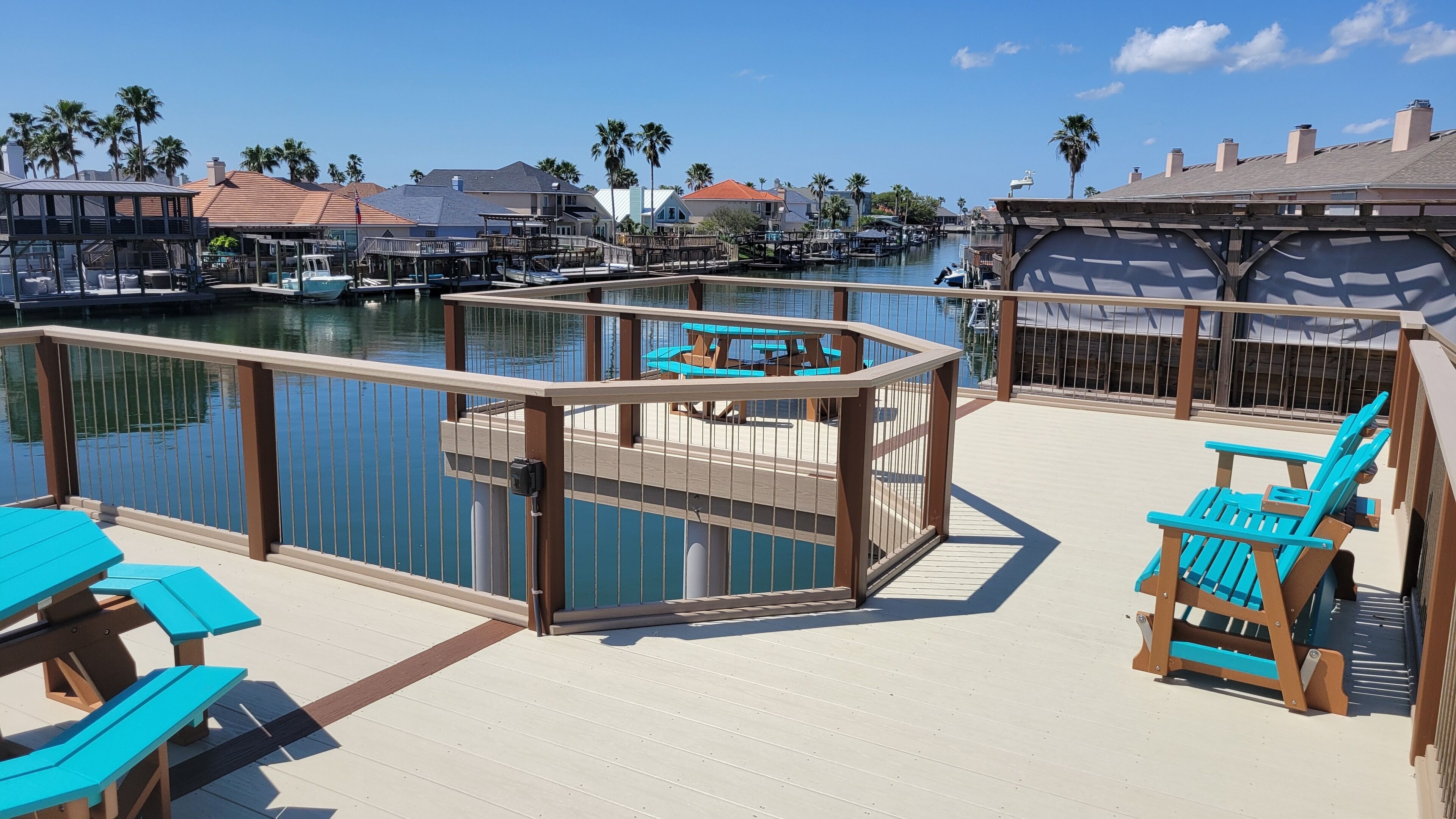 Un port de plaisance avec des bateaux amarrés, une terrasse en bois avec des chaises et des tables bleues, et une vue dégagée sur l’eau et les bâtiments environnants.