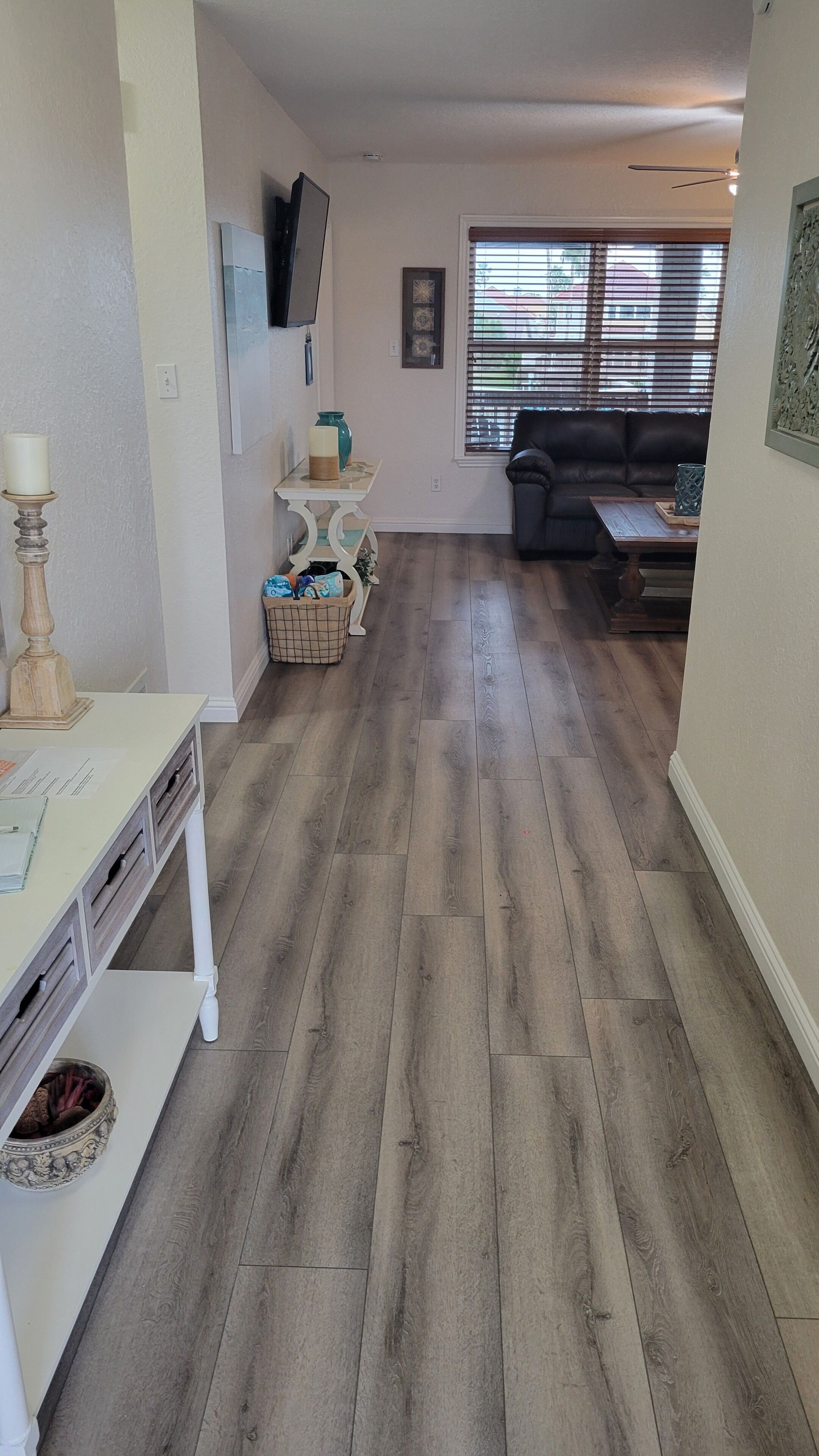 A spacious living area with hardwood flooring, a white entertainment unit, a black leather sofa, and a window with blinds.