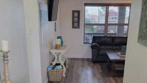 Amplia sala de estar con pisos de madera, un mueble blanco para entretenimiento, un sofá de cuero negro y una ventana con persianas.