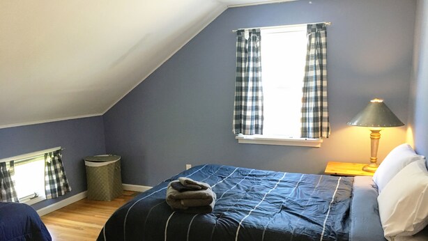 A bedroom with a sloped ceiling, a bed with a blue bedspread, a window with plaid curtains, a bedside lamp, and a trash can.