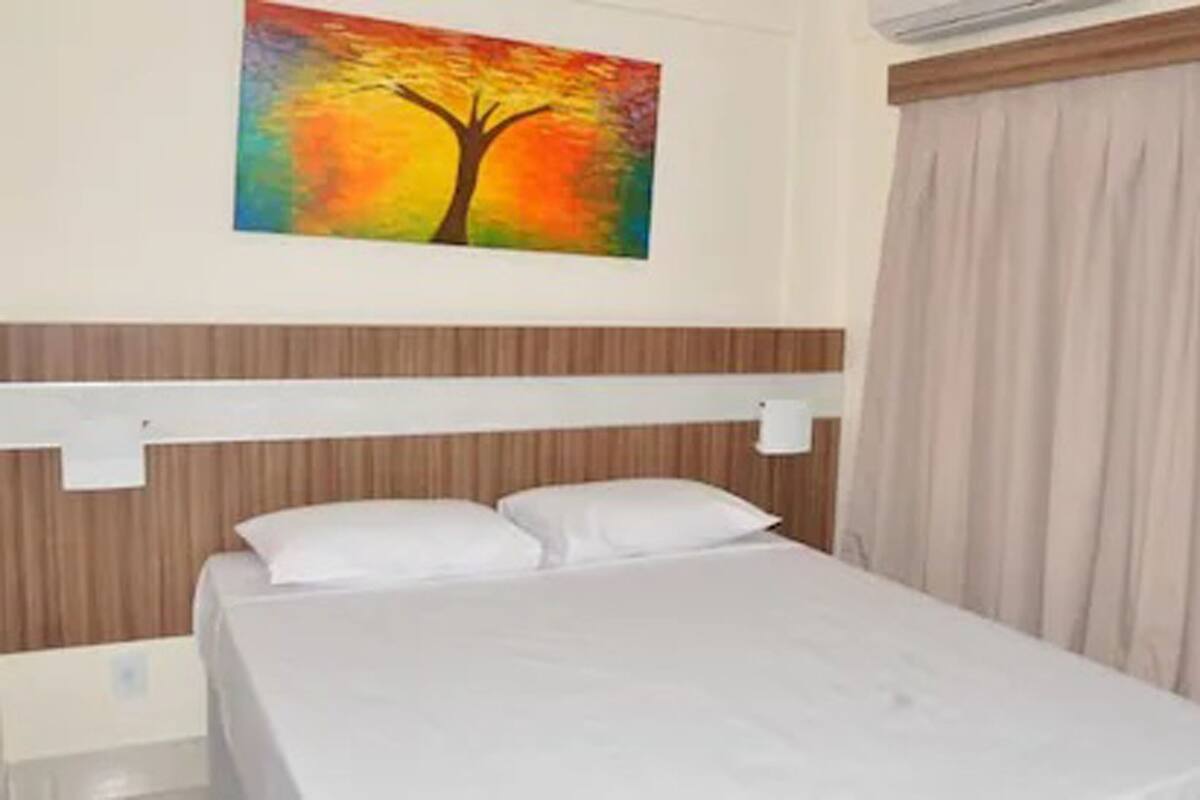 Habitación de hotel con cama, cabecera, un cuadro de un árbol y una ventana con cortinas.