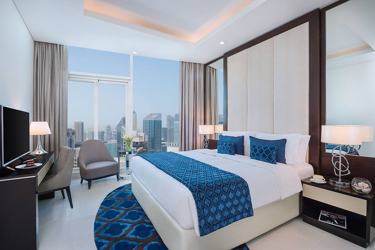 Una habitación de hotel moderna con una cama grande, dos mesitas de noche, un escritorio con silla y un ventanal con vistas a la ciudad.