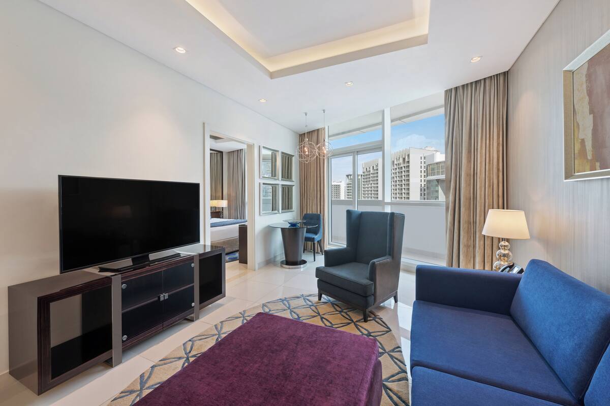 Una habitación de hotel moderna con televisor de pantalla plana, sofá, sillón, una mesita y ventana con vistas a la ciudad.
