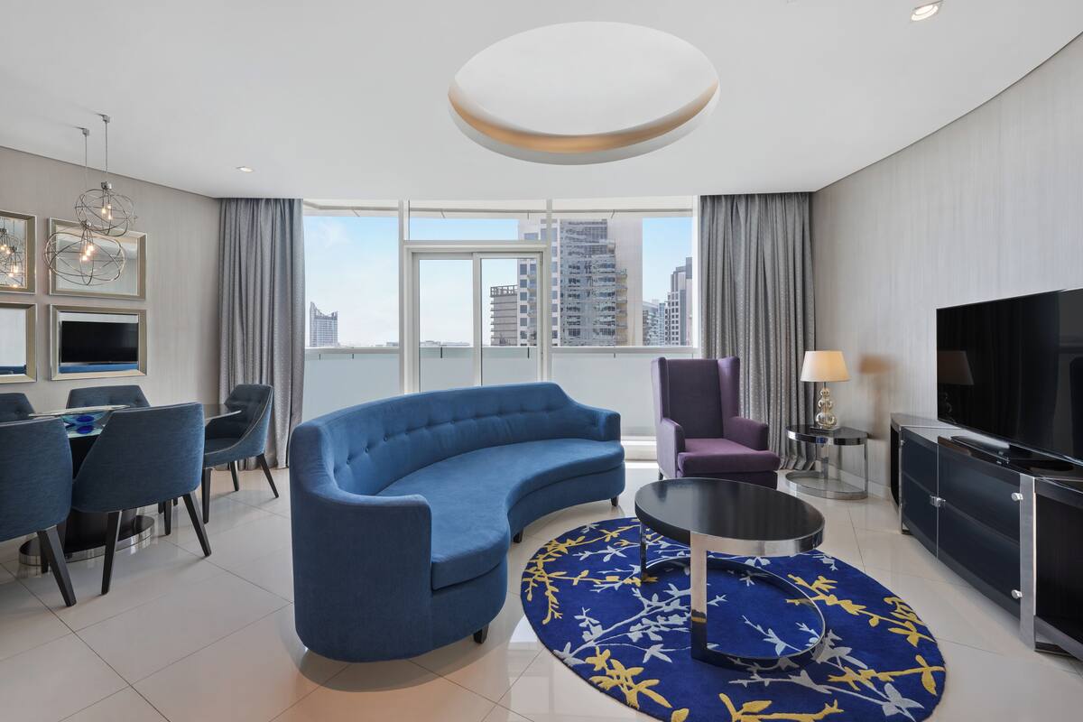 Una habitación de hotel moderna con un sofá azul curvo, un área de comedor con mesa y sillas, un televisor y un ventanal con vistas a la ciudad.