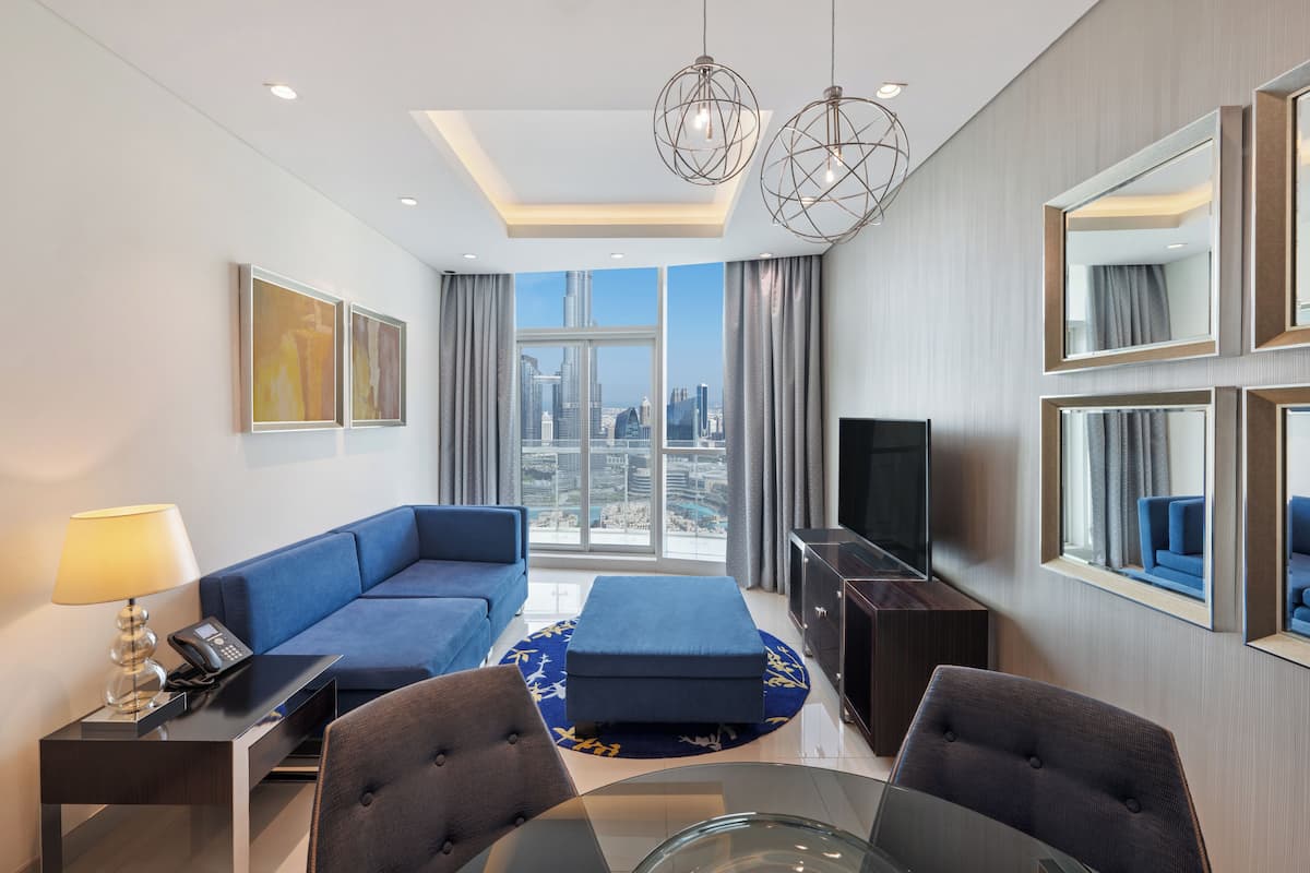 Habitación de hotel moderna con un ventanal grande que ofrece vistas a la ciudad, un sofá, un televisor y una zona de comedor con una mesa de cristal.