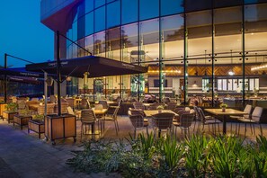 An outdoor seating area with tables and chairs, a large umbrella, and a building with glass windows in the background. - DAMAC Maison Distinction (Dubai)