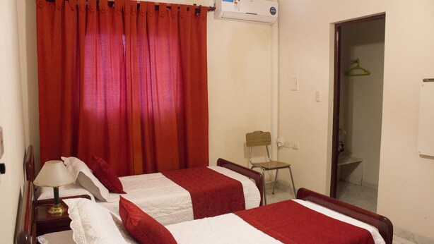 Habitación de hotel con dos camas, ropa de cama roja y blanca, una mesita de noche de madera con una lámpara, una silla y un equipo de aire acondicionado.