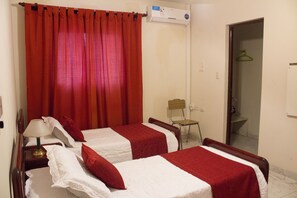 A hotel room with two beds, red and white bedding, a wooden nightstand with a lamp, a chair, and an air conditioning unit. - Departamento Urquiza (Salta)