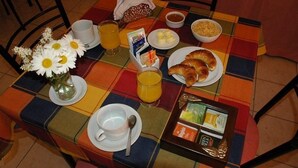 Una mesa de desayuno con mantel a cuadros, un florero de margaritas, cruasanes, jugo de naranja, cereal, mantequilla y una bandeja de pasteles.