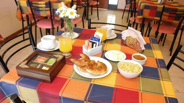 Una mesa de desayuno con mantel a cuadros, un jarrón con flores, una bandeja con cafetera, un croissant, un plato con huevos, un bol de fruta y un vaso de jugo de naranja.