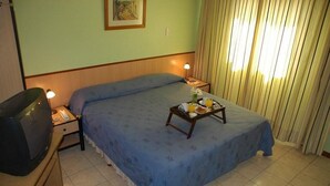 Habitación de hotel con una cama individual, una mesita de noche con lámpara, una pequeña mesa con una bandeja de desayuno y un televisor sobre un mueble.