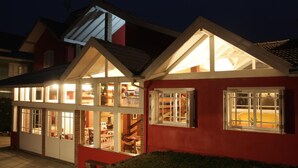 A two-story house with a red exterior and white trim, featuring large windows and a visible interior with furniture. - Aardvark Inn Pousada (Gramado)