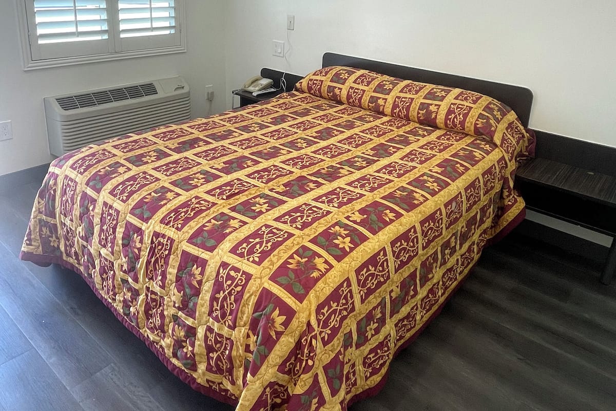 A bedroom with a large bed, a bedside table, a nightstand, a window with blinds, and a wall-mounted air conditioner.