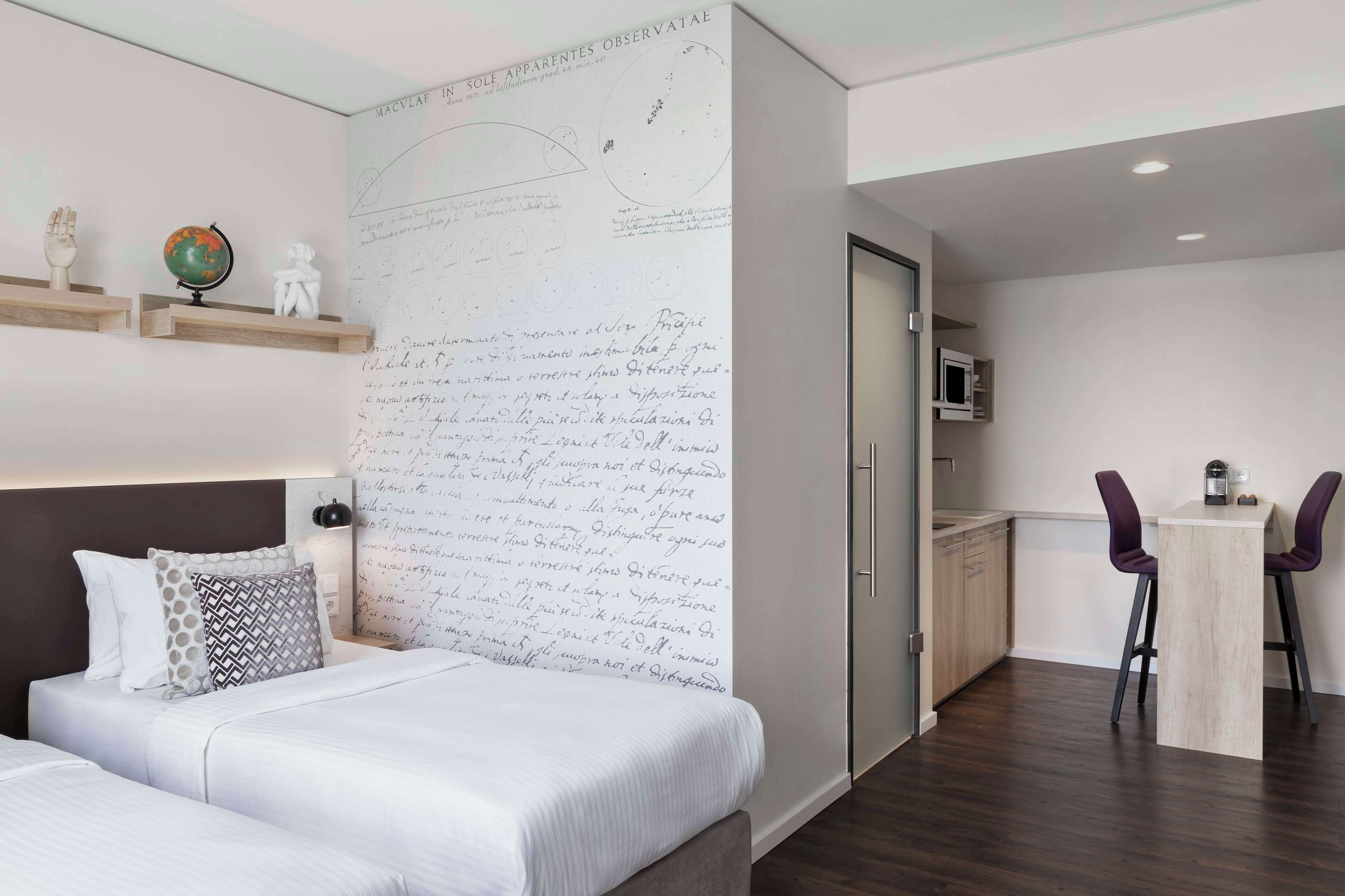 Una habitación de hotel moderna con una gran pared llena de notas escritas a mano, una cama con almohadas estampadas, un globo terráqueo y una pequeña estatua en un estante.