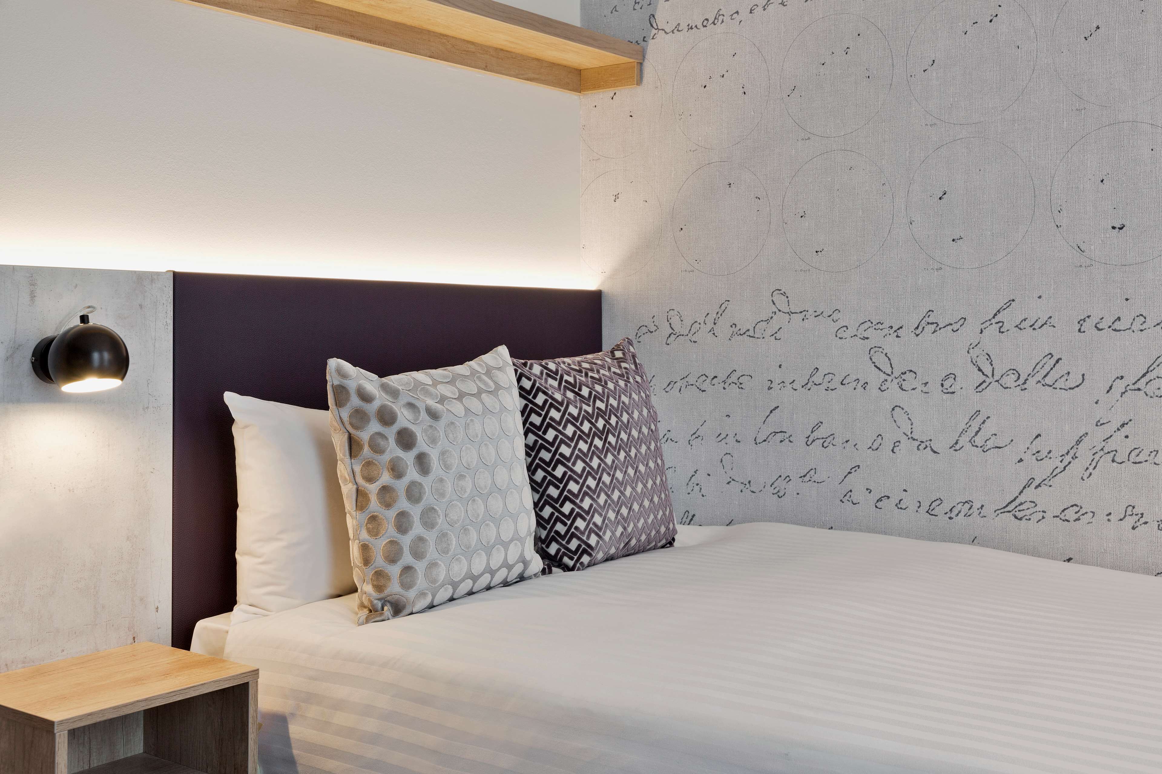 Quarto moderno com cama, duas almofadas estampadas, uma mesa de cabeceira e uma parede com padrões circulares.