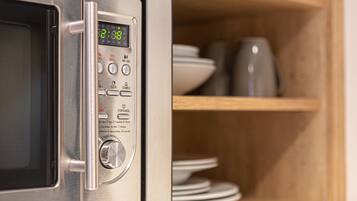 A microwave with a digital display and control panel, placed next to a wooden shelf with plates and a coffee maker.