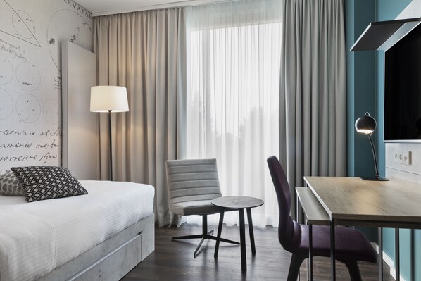 Habitación de hotel con cama, una silla, una mesita, una lámpara y vistas a través de cortinas translúcidas.