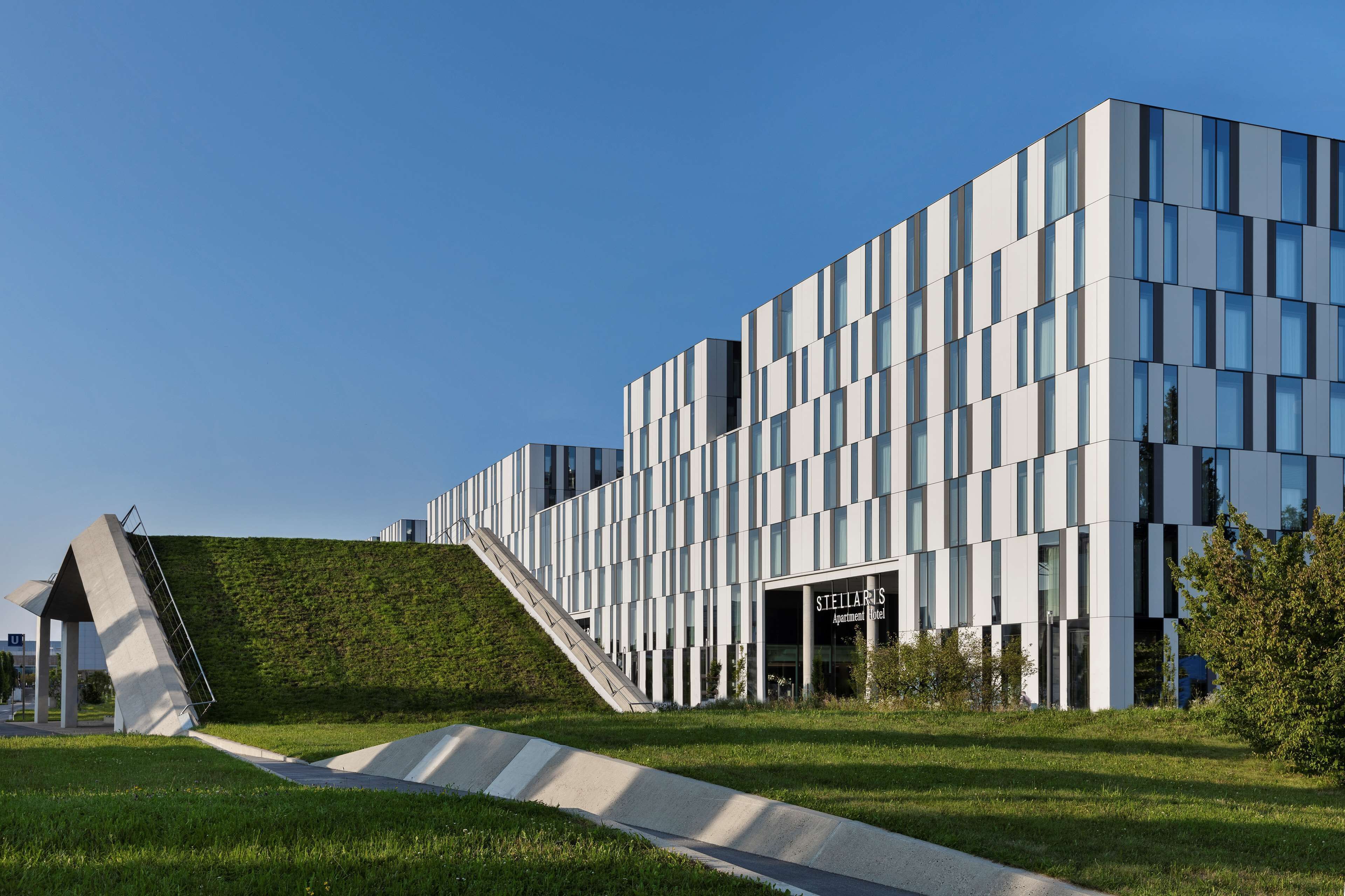 Ein modernes Gebäude mit einer markanten Fassade, einem geneigten Dach und einem gut sichtbaren Eingang, der „Stellaris Apartments Hotel“ ausweist.