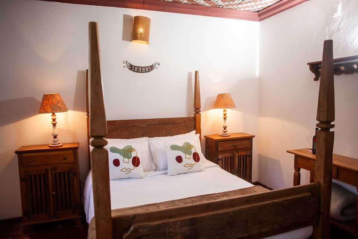 A four-poster bed with white bedding and two decorative pillows, flanked by wooden nightstands with lamps, in a room with a wooden ceiling and a wall-mounted light fixture.