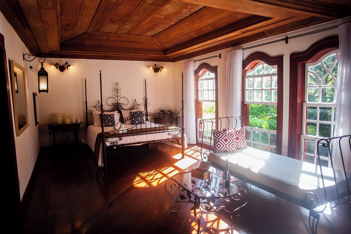 A room with a wooden ceiling, a bed with a metal frame, a bench, and a window with a view of greenery.