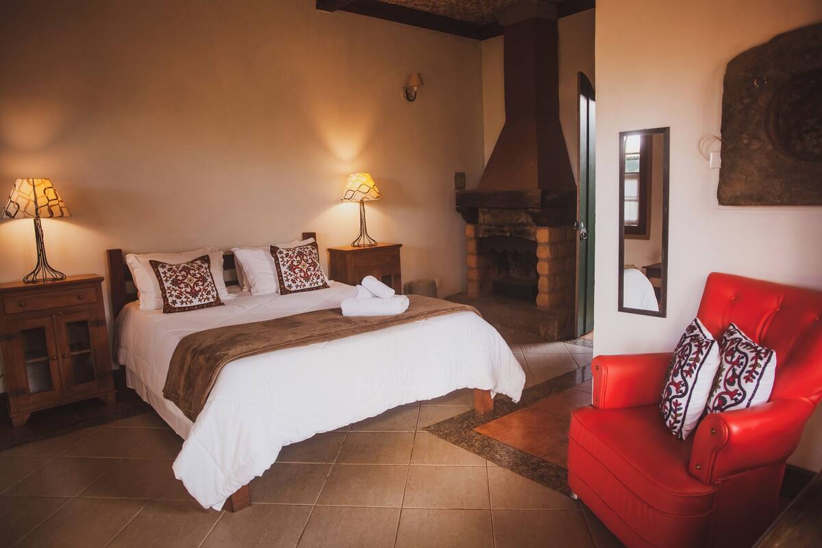 A bedroom with a large bed, two bedside tables with lamps, a red armchair, and a stone fireplace.