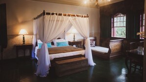 A four-poster canopy bed with white drapes, two bedside tables with lamps, a wooden bench, and a window with a view of greenery.