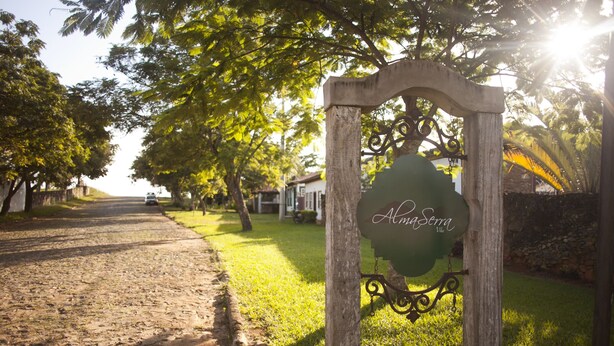 A rustic wooden sign with the name "AlmaSera" in a rural setting with a cobblestone path and lush greenery.
