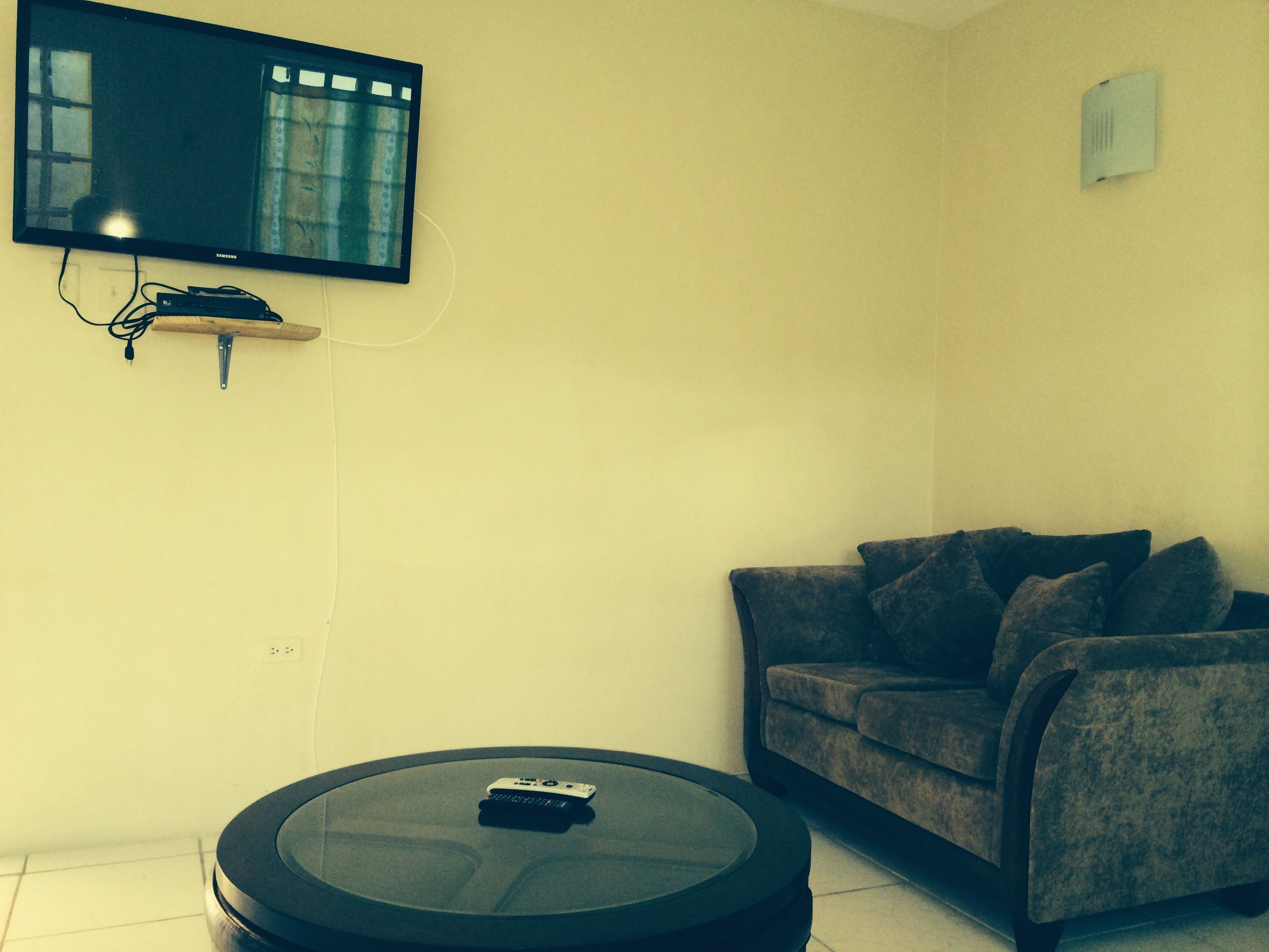 Uma sala de estar com televisão de tela plana na parede, uma mesa de centro redonda com um controle remoto e um sofá cinza.
