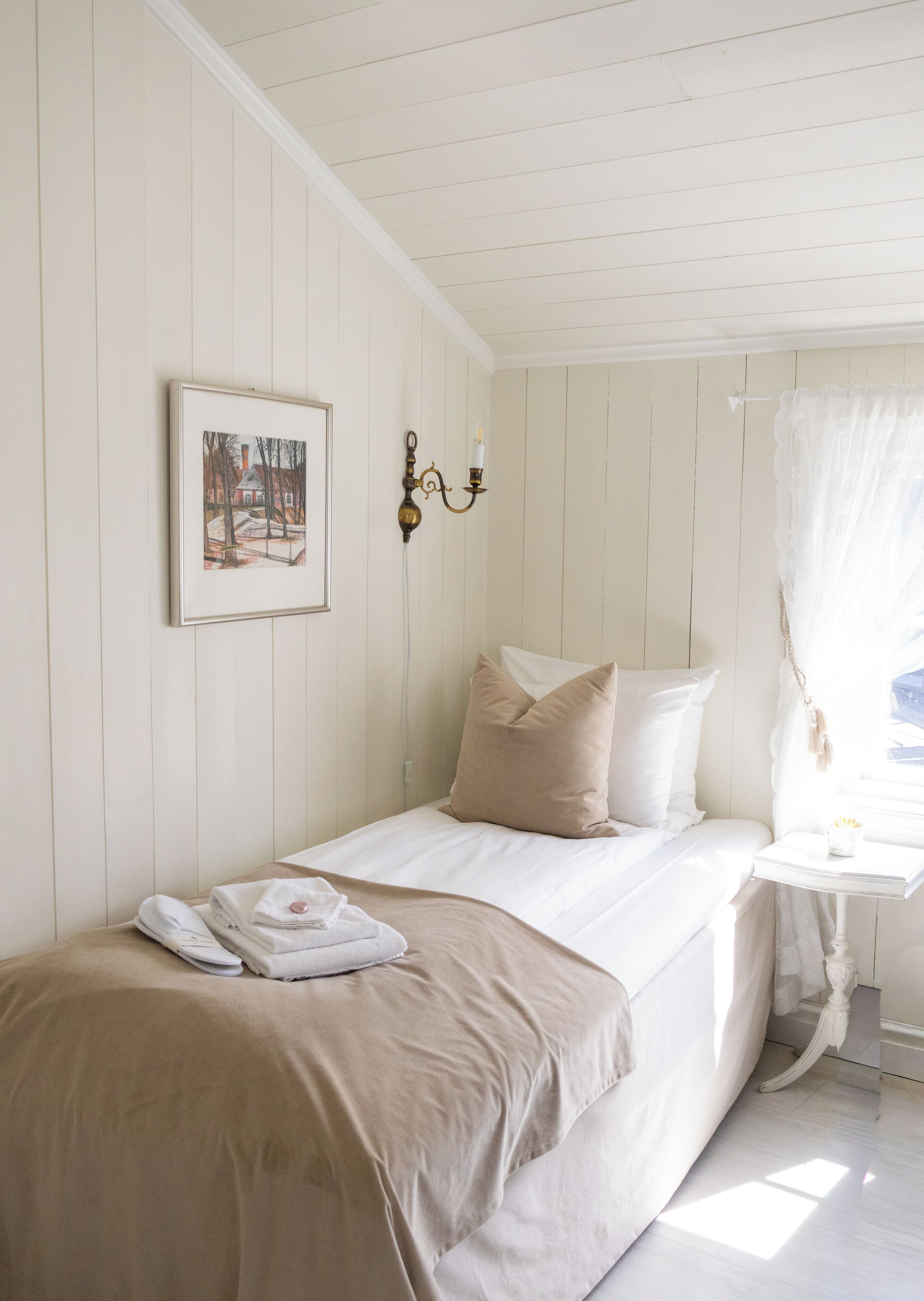 A single bed with a beige blanket, a framed picture on the wall, and a small table with a candle.