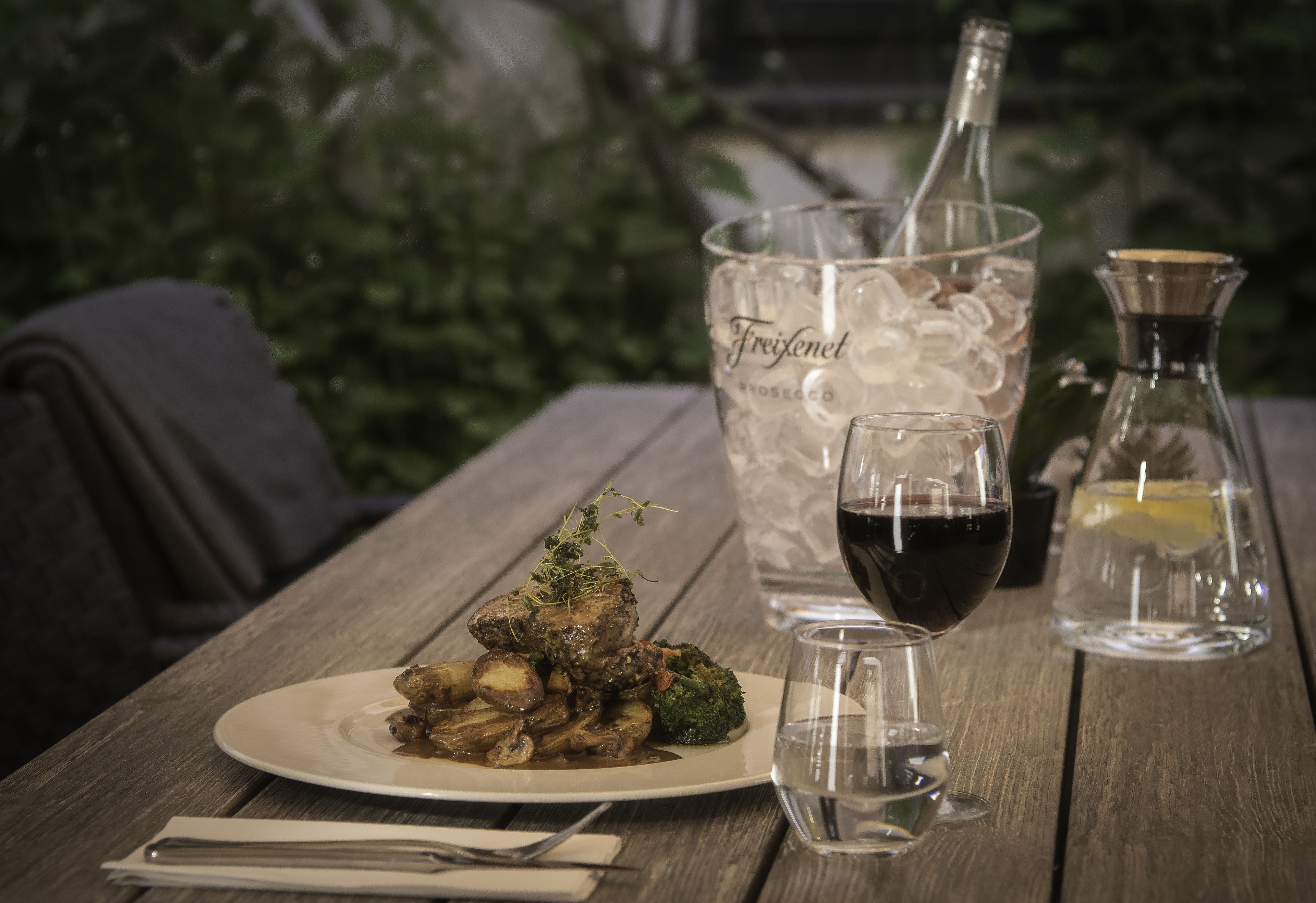 A wooden table with a plate of food, a glass of red wine, and a bottle of Freixenet rosé.